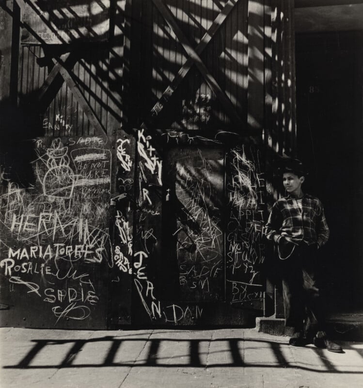 A young boy in a plaid shirt and hat stands against a graffiti-covered metal door, cast in the shadows of a fire escape.
