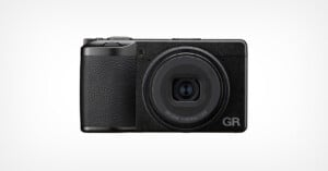 A black compact digital camera with a textured grip is shown facing forward against a plain white background. The lens is centered, and "GR" is visible on the lower right corner of the camera body.
