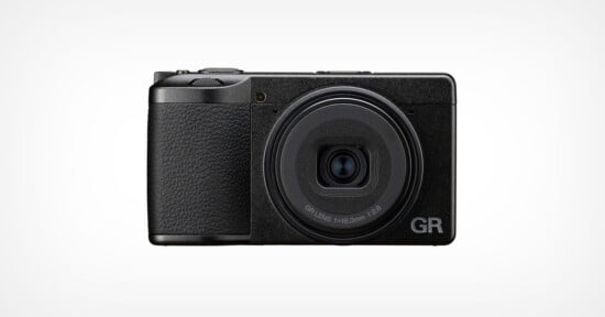 A black compact digital camera with a textured grip is shown facing forward against a plain white background. The lens is centered, and "GR" is visible on the lower right corner of the camera body.