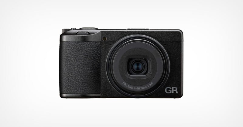 A black compact digital camera with a textured grip is shown facing forward against a plain white background. The lens is centered, and "GR" is visible on the lower right corner of the camera body.