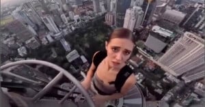 A woman with a backpack stands high on a metal structure, looking up at the camera with a concerned expression, while a cityscape with tall buildings spreads out far below her.