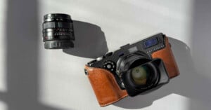 A Hasselblad camera with a brown leather case sits on a gray surface beside a detached camera lens. Shadows from a window fall across the scene.