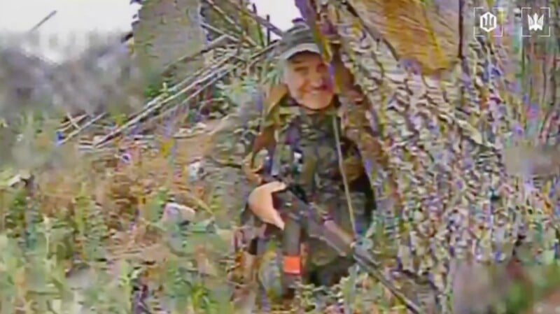 A person in camouflage clothing holding a rifle stands behind a tree in an outdoor, wooded area. The image appears slightly pixelated, and faint logos are visible in the top right corner.