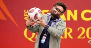 A man wearing glasses and a gray hoodie holds a soccer ball in both hands, standing in front of a red wall with large yellow text. He has an event badge and a smartwatch, and appears to be making a playful facial expression.