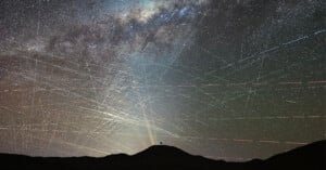 A dark silhouette of a hill is seen under a sky filled with bright star trails and the Milky Way, crisscrossed by numerous streaks of light from satellites and meteors.