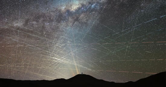 A dark silhouette of a hill is seen under a sky filled with bright star trails and the Milky Way, crisscrossed by numerous streaks of light from satellites and meteors.