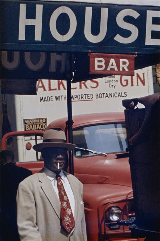 A man in a fedora and light-colored suit with a patterned tie stands in front of a red truck and a sign for a bar.