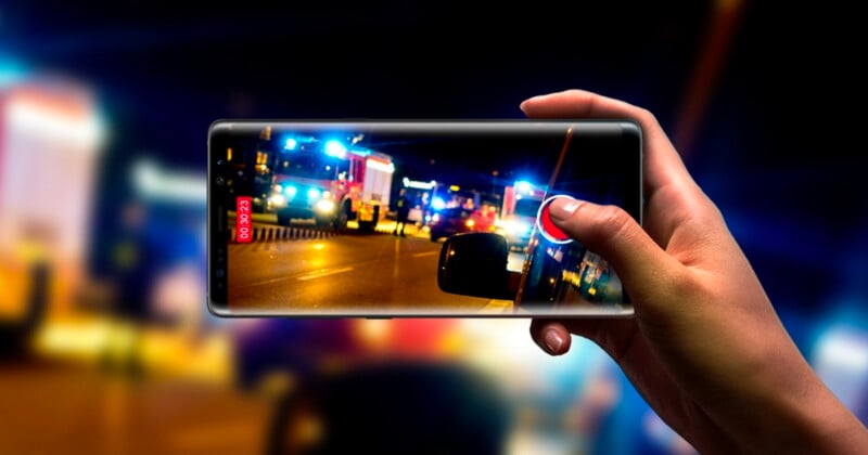 A hand holds a smartphone, capturing a photo of emergency vehicles with flashing lights at night, possibly at an accident scene. The phone screen shows a clearer image than the blurred background.
