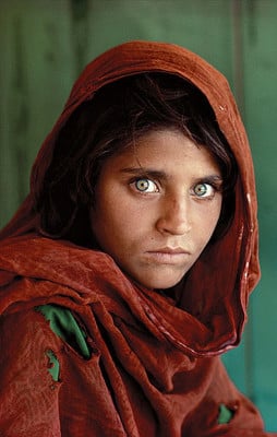 A young girl with striking green eyes and dark hair wears a tattered, dark red headscarf against a green background.