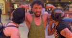 A smiling man in a green tank top flexes his arms while two people on either side pretend to kiss his biceps. They are outside at night under bright lights.