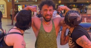 A smiling man in a green tank top flexes his arms while two people on either side pretend to kiss his biceps. They are outside at night under bright lights.