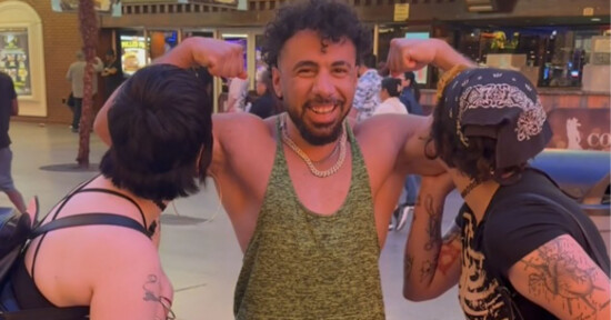 A smiling man in a green tank top flexes his arms while two people on either side pretend to kiss his biceps. They are outside at night under bright lights.