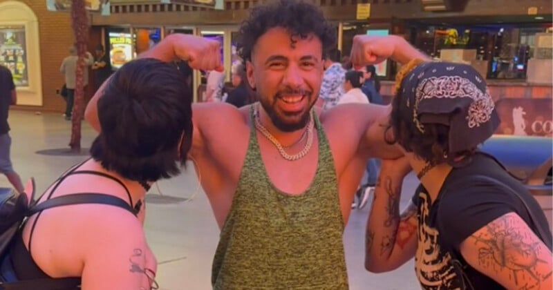 A smiling man in a green tank top flexes his arms while two people on either side pretend to kiss his biceps. They are outside at night under bright lights.