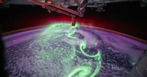 A view from the International Space Station shows swirling green and purple auroras above Earth’s atmosphere, with parts of the space station visible in the foreground.