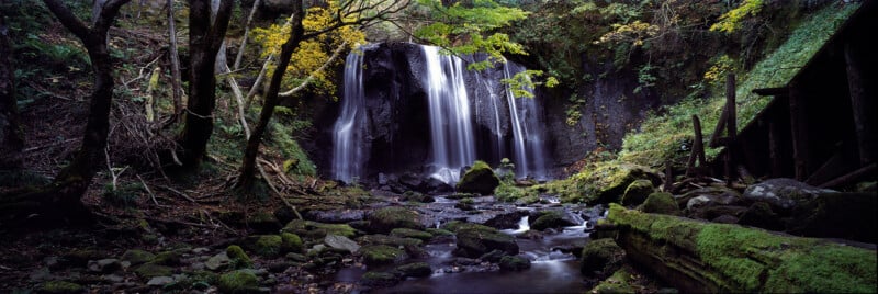 A wide waterfall cascades over a dark, rocky cliff into a mossy, boulder-strewn stream surrounded by a dense forest.