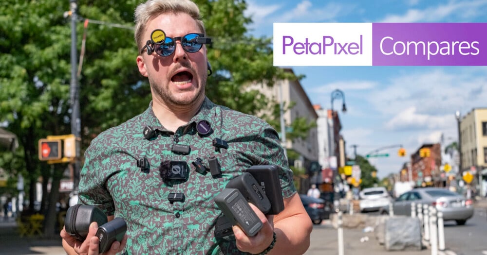 A man with light hair and sunglasses, wearing multiple cameras and holding camera gear, stands on a sunny city street. The "PetaPixel Compares" logo is overlaid on the image.
