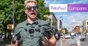 A man with light hair and sunglasses, wearing multiple cameras and holding camera gear, stands on a sunny city street. The "PetaPixel Compares" logo is overlaid on the image.