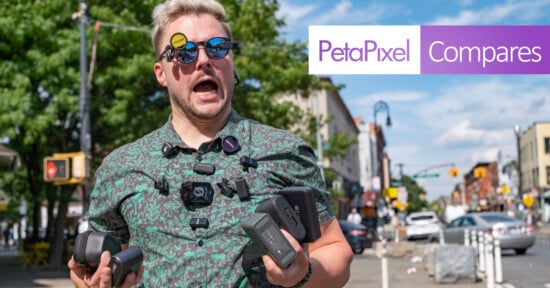 A man with light hair and sunglasses, wearing multiple cameras and holding camera gear, stands on a sunny city street. The "PetaPixel Compares" logo is overlaid on the image.