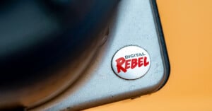Close-up of a corner of a silver camera with a round button labeled "DIGITAL REBEL" in black and red text. The background is a solid orange color.