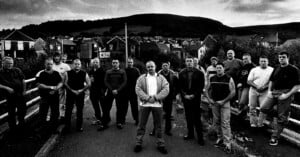 A group of men stand on a road in front of houses and hills, posed together in an intense, serious manner. The photo is in black and white, giving it a dramatic and moody atmosphere.