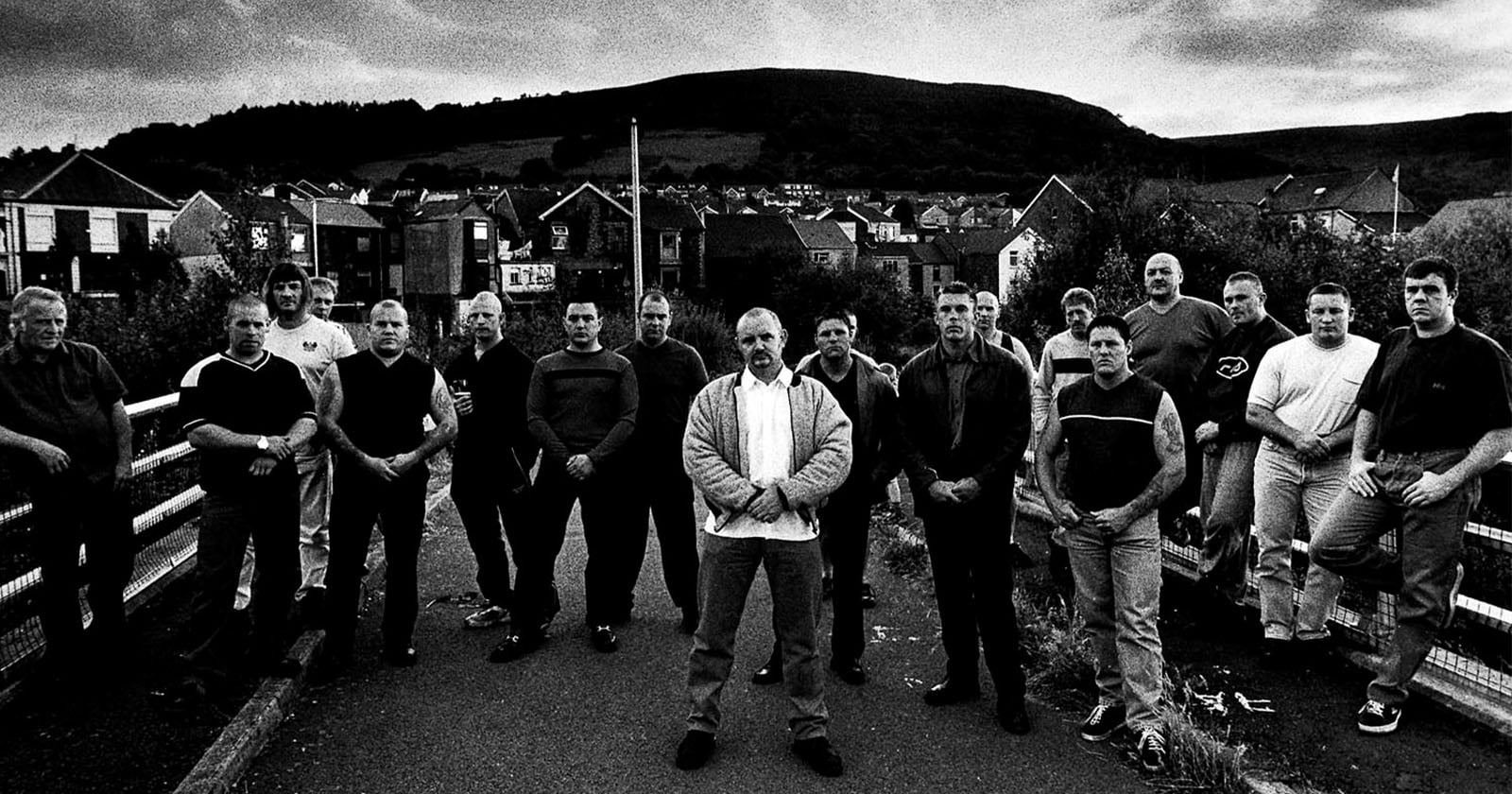 A group of men stand on a road in front of houses and hills, posed together in an intense, serious manner. The photo is in black and white, giving it a dramatic and moody atmosphere.