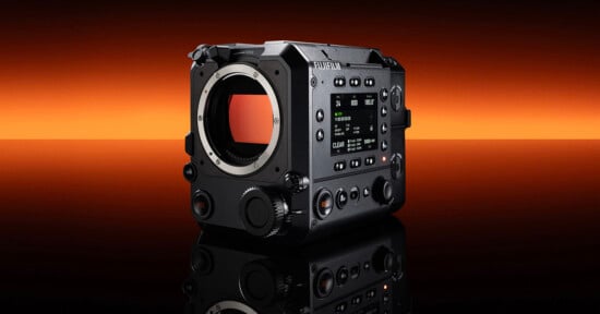 A black Fujifilm digital cinema camera body is displayed on a reflective surface with an orange to black gradient background. The camera features multiple dials, buttons, and a digital screen on its side.