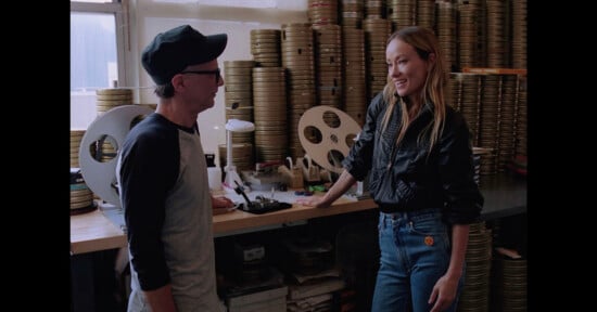 A man and woman talk in a room filled with film reels. The man wears glasses, a cap, and a baseball shirt, while the woman wears a black jacket and jeans, leaning against a desk with editing equipment.