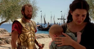 A man in golden armor stands near a woman holding a baby, with ships anchored by the shore and olive trees in the background.