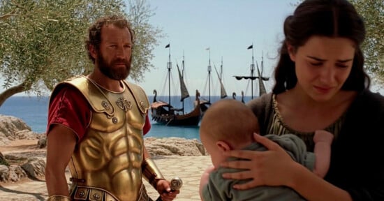 A man in golden armor stands near a woman holding a baby, with ships anchored by the shore and olive trees in the background.
