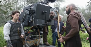 Actors in medieval costumes stand near a large film camera outdoors on a grassy set. Crew members adjust equipment while one actor holds a bow and another has arrows on his back.