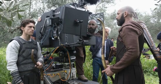 Actors in medieval costumes stand near a large film camera outdoors on a grassy set. Crew members adjust equipment while one actor holds a bow and another has arrows on his back.