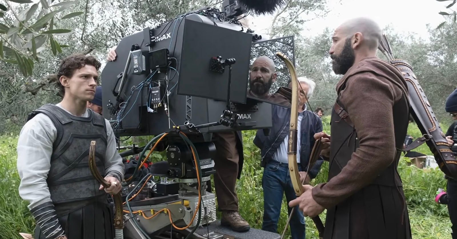 Actors in medieval costumes stand near a large film camera outdoors on a grassy set. Crew members adjust equipment while one actor holds a bow and another has arrows on his back.