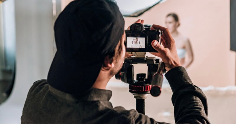 A photographer wearing a black baseball cap looks through the viewfinder of a camera mounted on a tripod, focusing on a model in the background.