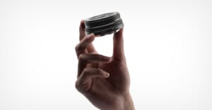 A hand holds a small camera lens delicately between the thumb and index finger against a plain white background.