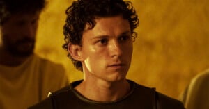 A young man with curly brown hair wearing a dark shirt and armor looks ahead seriously; the background is yellow with a blurred figure behind him.