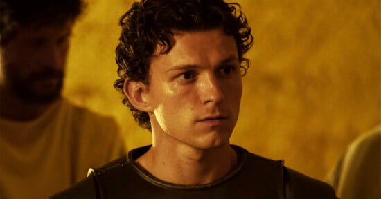 A young man with curly brown hair wearing a dark shirt and armor looks ahead seriously; the background is yellow with a blurred figure behind him.