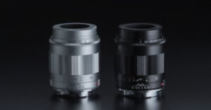 Two camera lenses, one silver and one black, are standing upright side by side on a reflective dark surface, both showing aperture and focus markings.