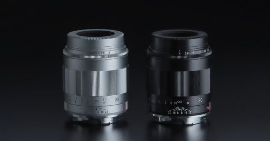 Two camera lenses, one silver and one black, are standing upright side by side on a reflective dark surface, both showing aperture and focus markings.