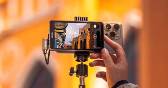 A hand holds a smartphone mounted on a tripod, with a Viltrox external monitor attached to the phone displaying a live camera feed of a street scene.