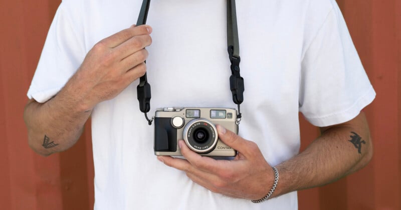 A person in a white t-shirt holds a vintage camera by its strap. Their left arm has a tattoo, and they wear a silver bracelet. The background is a red wall.
