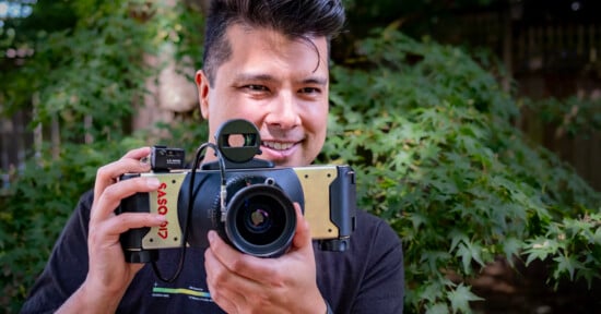 A smiling man holds a wide, metallic panoramic camera with a top-mounted viewfinder in front of his face against a background of green foliage.
