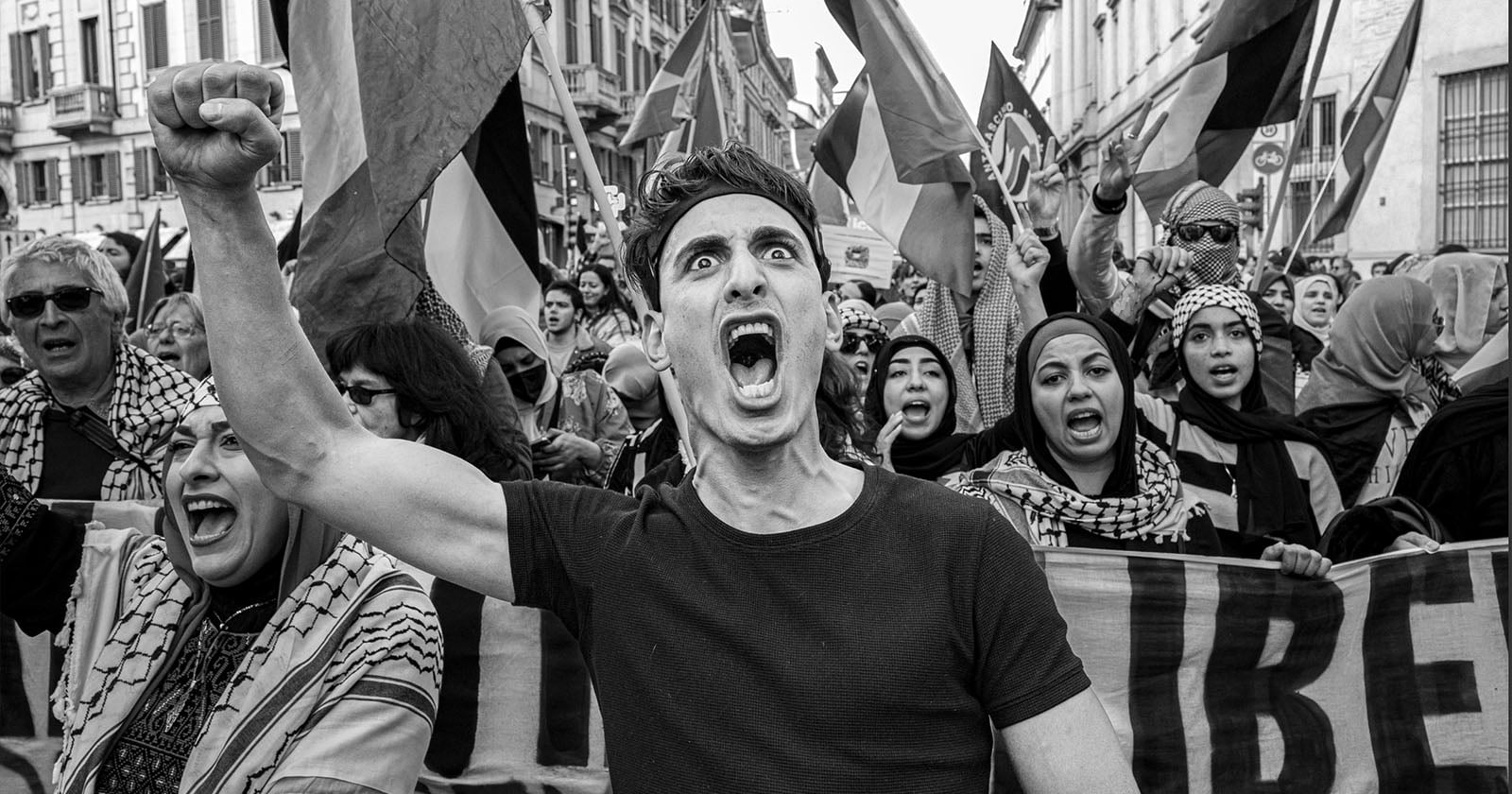 A man with a raised fist shouts passionately at the front of a large protest crowd, many holding flags and banners, with expressions of determination and emotion.