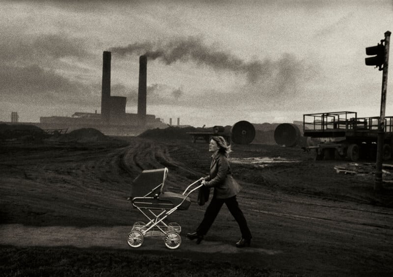 A woman pushes a baby stroller across a muddy industrial landscape with smoke billowing from factory chimneys in the background. The scene is dark and gritty, evoking a sense of hardship.
