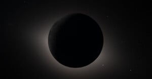 A dark celestial body, likely a moon or planet, is silhouetted against a faint, glowing halo of light with a starry background in space. The object's surface features are barely visible.
