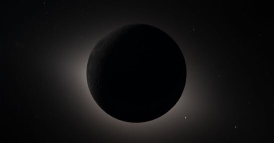 A dark celestial body, likely a moon or planet, is silhouetted against a faint, glowing halo of light with a starry background in space. The object's surface features are barely visible.