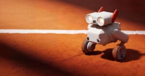 A small, white, robot with two wheels and two red antennae stands on a clay tennis court near a white line, illuminated by sunlight.