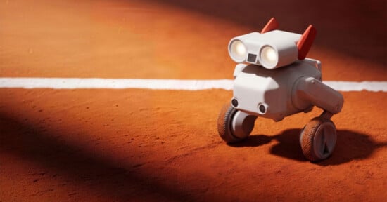 A small, white, robot with two wheels and two red antennae stands on a clay tennis court near a white line, illuminated by sunlight.