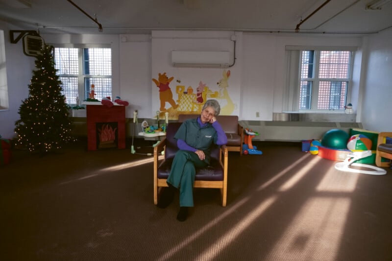 An older woman sits on a chair in a playroom decorated with a Christmas tree and Winnie the Pooh mural, surrounded by scattered toys and exercise balls. Sunlight streams through the windows.