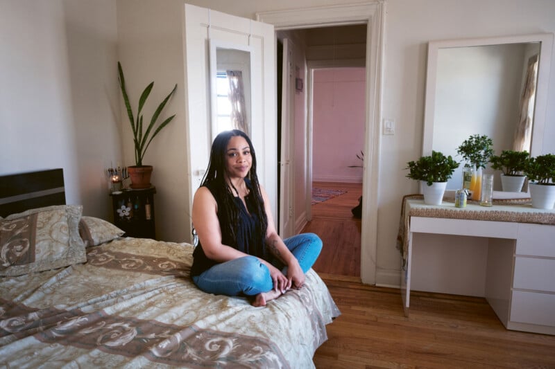 A woman with long hair sits cross-legged on a bed in a tidy bedroom with plants, a vanity, and neutral decor. A door to another room is open in the background.
