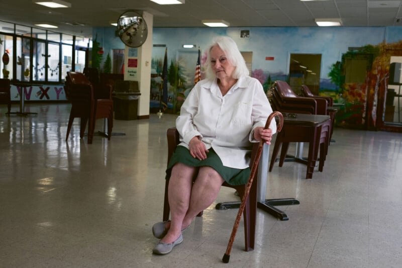 An older woman with white hair sits on a chair in an empty room, holding a cane. She wears a white blouse, green skirt, and gray shoes. Stacked chairs, tables, and a mural are visible in the background.